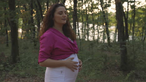 Mujer-Embarazada-Mirando-Alrededor-Del-Bosque-Y-Hacia-Abajo-En-El-Vientre-Mientras-Camina-Al-Atardecer