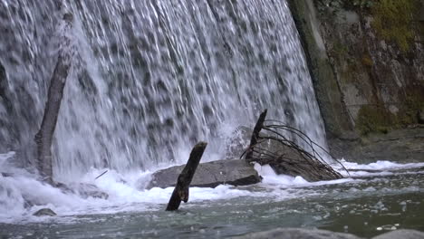 Schweizer-Fluss,-Der-Nach-Einem-Wasserfall,-Einigen-Baumstämmen-Und-Kieseln-Schäumt