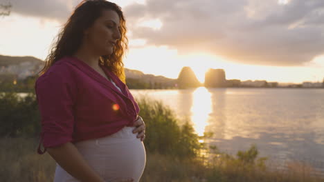 Schwangere-Mutter-Tief-In-Gedanken-Versunken-Bei-Sonnenuntergang-Lächelnd-Und-Bauch-Streichelnd-Am-Wasser