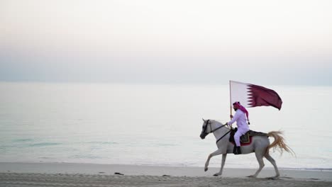 Ein-Ritter,-Der-Auf-Einem-Pferd-Reitet-Und-Die-Katar-flagge-In-Der-Nähe-Des-Meeres-Hält