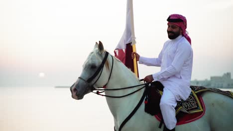 Ein-Ritter,-Der-Auf-Einem-Pferd-Reitet,-Das-Die-Katar-flagge-Nahe-Dem-Meer-Hält