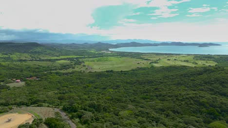 Aerial-view-of-a-landscape-that-has-mountains-far-away,-with-green-pastures,-grasslands,-trees,-and-beaches-during-a-sunny-day-while-birds-fly-through-the-shot