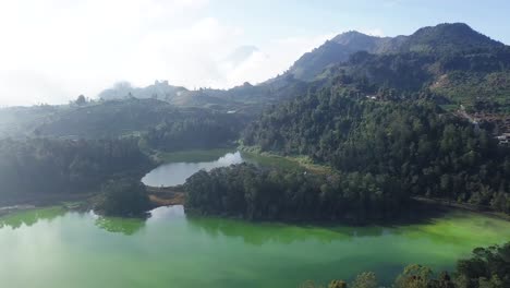 Drone-shot-flight-over-natural-TELAGA-WARNA-Lake-surrounded-by-green-forest-trees-growing-on-mountain-during-sunny-day