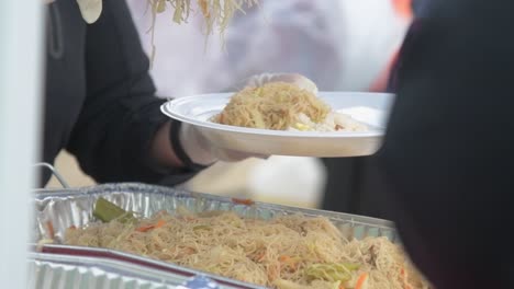 Person-serving-fresh-hot-noodles-on-white-plastic-plate-with-tongs,-outdoor-buffet