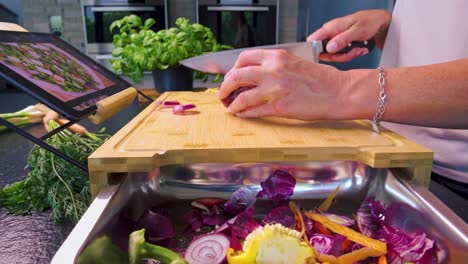 Close-up-panning-shot-of-a-women-cutting-vegetables-on-a-cutting-board-with-a-recipe-on-a-tablet-in-a-modern-kitchen-and-fresh-herbs-and-vegetables