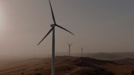 Close-up-tracking-aerial-shot-of-several-wind-turbines-at-sunset-or-sunrise-on-a-dusty-foggy-day