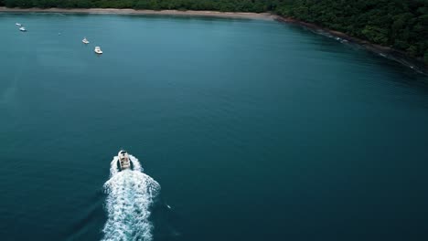 Luftdrohnenaufnahme-Eines-Segelboots,-Das-Durch-Einen-Blauen-Golf-In-Richtung-Ufer-Segelt,-Mit-Mehr-Booten-Im-Hintergrund-Und-Vegetaino-Am-Ufer