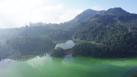 Drone-shot-flight-over-natural-TELAGA-WARNA-Lake-surrounded-by-green-forest-trees-growing-on-mountain-during-sunny-day---Central-Java,Indonesia