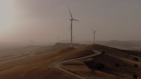 Tracking-aerial-shot-of-a-winding-road-leading-towards-several-wind-turbines-at-sunset-or-sunrise-on-a-dusty-foggy-day