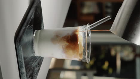 Vertical-Video-home-barista-making-iced-latte-pouring-espresso-shot-into-glass-drinking-cup-containing-milk-with-glass-straw-in-slow-motion-close-up
