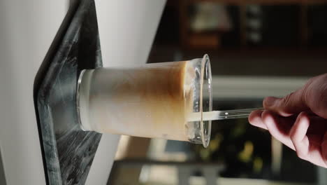 Vertikaler-Video-Heimbarista,-Der-Kaffee-Und-Milch-Rührt,-Um-Eislatte-In-Einem-Glastrinkbecher-Mit-Glasstrohhalm-In-Zeitlupe-Aus-Nächster-Nähe-Zuzubereiten