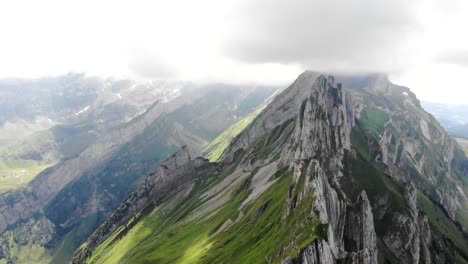Aerial-flyover-over-and-away-from-the-cliffs-of-Schafler-ridge-in-Appenzell,-Switzerland-with-cliffs,-mountain-peaks-and-lush-summer-green-mountainside-in-view