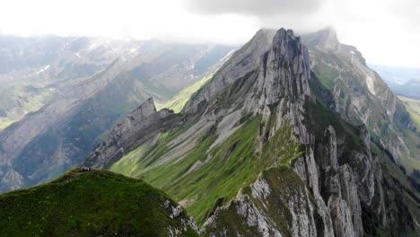 Aerial-flyover-over-the-side-of-Schafler-ridge-in-Appenzell,-Switzerland-with-cliffs,-mountain-peaks-and-lush-summer-green-mountainside-in-view