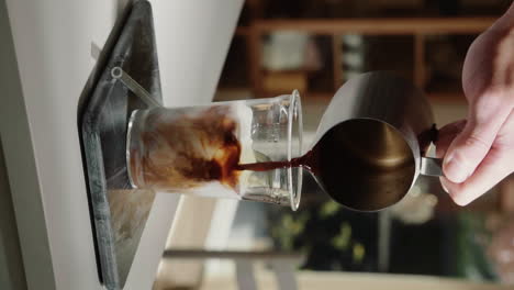 Vertical-Video-home-barista-making-iced-latte-pouring-espresso-shot-into-glass-drinking-cup-containing-milk-with-glass-straw-in-slow-motion-close-up-1