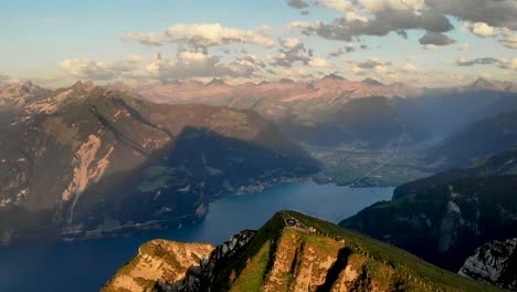 Aerial-flyover-away-from-the-summit-of-Niederbauen-Chulm-revealing-the-peak's-cliffs-with-a-view-of-Uri-and-Lake-Lucerne-fjords-in-the-background-on-a-summer-evening-in-the-Swiss-Alps