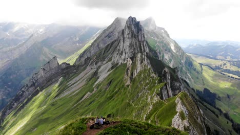 Sobrevuelo-Aéreo-Sobre-Los-Dos-Excursionistas-Disfrutando-De-La-Vista-De-La-Cresta-Schafler-En-Appenzell,-Suiza-Con-Acantilados-Y-Picos-Montañosos-A-La-Vista