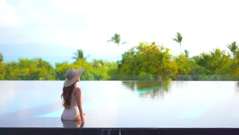Back-View-of-Sexy-Woman-in-Swimsuit-and-Hat-in-Infinity-Pool-With-Beautiful-View-of-Tropical-Island-and-Ocean-Horizon