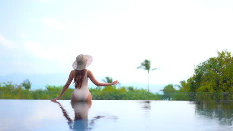 Back-View-of-Lonely-Sexy-Woman-Sitting-on-Infinity-Pool-Edge-With-Stunning-View-of-Tropical-Island-and-Sea-Horizon