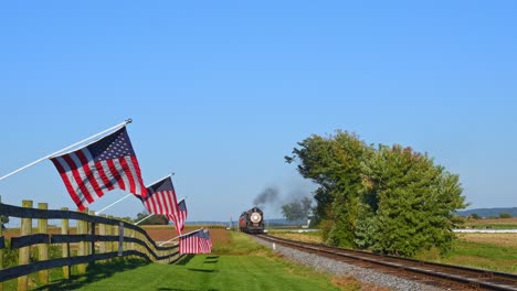 A-View-of-a-Line-of-Gently-Waving-American-Flags-on-a-Fence-by-Farmlands-as-a-Steam-Passenger-Train-Blowing-Smoke-Approaches-During-the-Golden-Hour