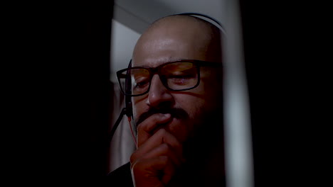 Bald-man-with-glasses,-moustache-and-headset-thinking-and-looking-at-a-computer-screen-that-flashes-bright-colors-while-he-holds-his-hand-in-his-mouth