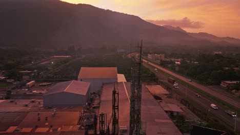Ascending-aerial-shot-of-industrial-factory-and-traffic-on-highway-during-golden-sunrise-behind-mountains---Villa-Altagracia,Dominican-Republic