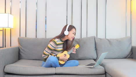 A-cute-young-woman-enjoys-sitting-on-the-couch-while-playing-a-yellow-ukulele-as-she-interacts-with-her-laptop