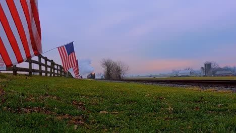 A-Low-Angle-View-of-a-Line-of-Gently-Waving-American-Flags-on-a-Fence-by-Farmlands-as-a-Steam-Passenger-Train-Blowing-Smoke-Approaches-During-the-Golden-Hour-1