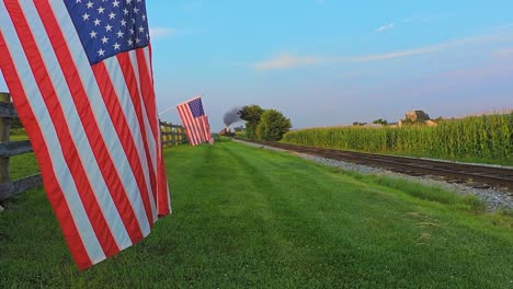 A-Low-Angle-View-of-a-Line-of-Gently-Waving-American-Flags-on-a-Fence-by-Farmlands-as-a-Steam-Passenger-Train-Blowing-Smoke-Approaches-During-the-Golden-Hour