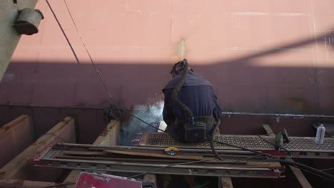 Man-in-protective-suit-sits-on-an-iron-beam-while-he-is-repairing-welding-an-old-ship