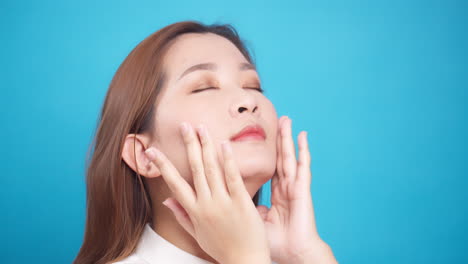Nahaufnahme-Einer-Perfekten-Hübschen-Asiatischen-Frau,-Die-Das-Gesicht-Sanft-Mit-Anti-Aging-Balsamcreme-Für-Wellness-Hautpflege-Auf-Blauem-Hintergrund-Reibt