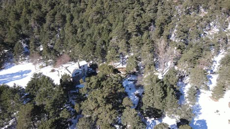 Flying-over-a-snowy-pine-forest-on-a-mountain-side-and-an-old-abandoned-shelter-building-in-the-wilderness