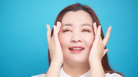Nahaufnahme-Einer-Perfekten-Hübschen-Asiatischen-Frau,-Die-Das-Gesicht-Sanft-Mit-Anti-Aging-Balsamcreme-Für-Wellness-Hautpflege-Auf-Blauem-Hintergrund-Reibt-1