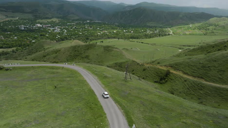 Aerial-View-Of-White-Vehicle-Driving-In-The-Road-Overlooking-The-Green-Hills,-Meadow,-And-Mountains-Near-Aspindza-In-Georgia