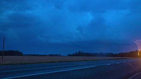 Timelapse-shot-of-cars-driving-along-rural-road-with-lightning-in-dark-clouds-along-the-background-during-evening-time