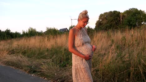 Slow-motion-shot-of-caucasian-pregnant-woman-in-summer-dress-touching-belly-and-walking-outdoors-between-grass-fiel-park-at-sunset