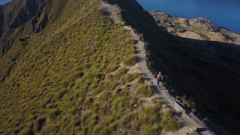 Tracking-aerial-shot-of-a-tall-blonde-woman-running-along-a-trail-to-the-top-of-a-mountain-and-then-stopping-and-gaining-her-balance