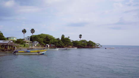 Vista-Aérea-Baja-De-La-Bahía-De-Pescadores-En-La-Playa-Del-Tesoro-Jamaica-Con-Barcos-De-Pesca-Flotando-En-La-Costa-Y-Elevándose-Sobre-El-Afloramiento-Rocoso-De-La-Tierra