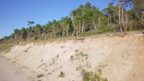 Aerial-view-of-Baltic-sea-beach-at-Jurkalne-on-a-sunny-day,-white-sand-cliff-damaged-by-waves,-coastal-erosion,-climate-changes,-wide-angle-ascending-drone-shot-moving-forward