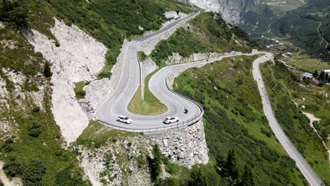 Tracking-panoramic-aerial-shot-of-two-white-cars-and-a-moto-taking-a-very-tight-curve-in-Grimselpass,-Switzerland