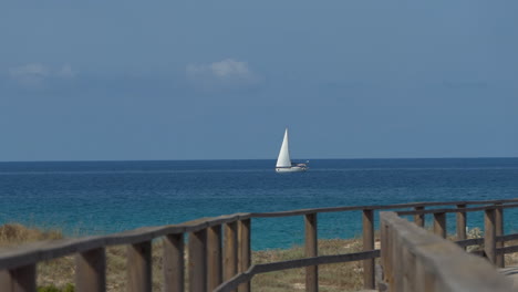 Small-white-sailboat-sailing-fast-on-the-azure-sea-near-the-coast,-clear-sky-with-few-clouds,-barriers-of-a-nice-wooden-path-in-the-foreground