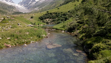 Luftbild,-Das-Sich-Durch-Einen-Bergsee-Mit-Türkisfarbenem,-Klarem-Wasser-Bewegt-Und-Im-Sommer-Einem-Fluss-Folgt,-Mit-Schneebedeckten-Bergen-Im-Hintergrund