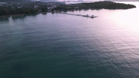 Drone-aerial-pan-up-over-tropical-blue-water-to-pier-during-sunrise-in-green-mountainside