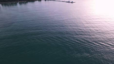 Drone-aerial-pan-up-over-tropical-blue-water-to-pier-during-sunrise