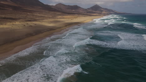 Fantastic-aerial-shot-over-the-sea,-seeing-the-mountains-and-the-waves-breaking-on-the-shore-of-Cofete-beach