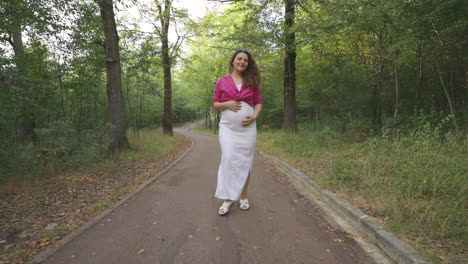 Happy-Pregnant-Woman-Expectant-Mother-Walking-Alone-in-Nature,-Attractive-Curly-Casually-Dressed-Mom-Turning-Around-Stroking-Caressing-Touching-Her-Belly-Smiling,-Childbearing-Love,-followed-by-camera