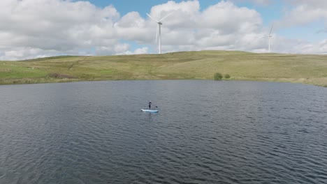 Close-Circling-Shot-Of-A-Individual-On-A-SUP-With-Windmills-Spinning-In-The-Background