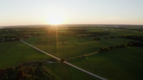 Aerial-drone-shot-over-rural-landscape-with-roads-surrounded-by-green-grasslands-on-bothside-in-Wisconsin,-USA-with-sunrise-over-horizon