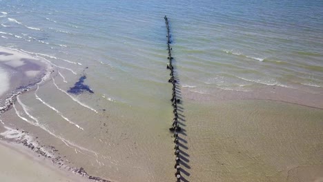 Aerial-birdseye-view-of-Baltic-sea-coast-on-a-sunny-day,-old-wooden-pier,-white-sand-coastline-damaged-by-waves,-coastal-erosion,-climate-changes,-wide-angle-drone-shot-moving-forward