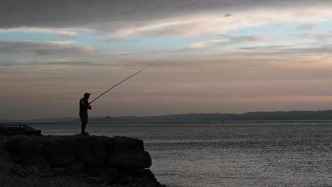 Old-man-fishing-from-a-rock-pier-on-the-lake-during-beautiful-summer-sunset