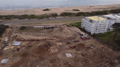 Aerial-view-of-construction-site-in-front-of-ocean-and-sandy-beach-of-Punta-del-Este---Cars-on-coastal-road-driving-along